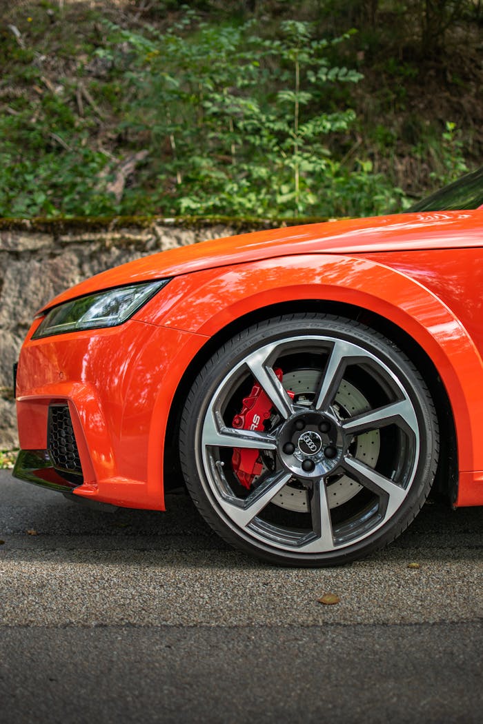 Detailed shot of a sleek orange car wheel on a sunlit road surrounded by greenery.
